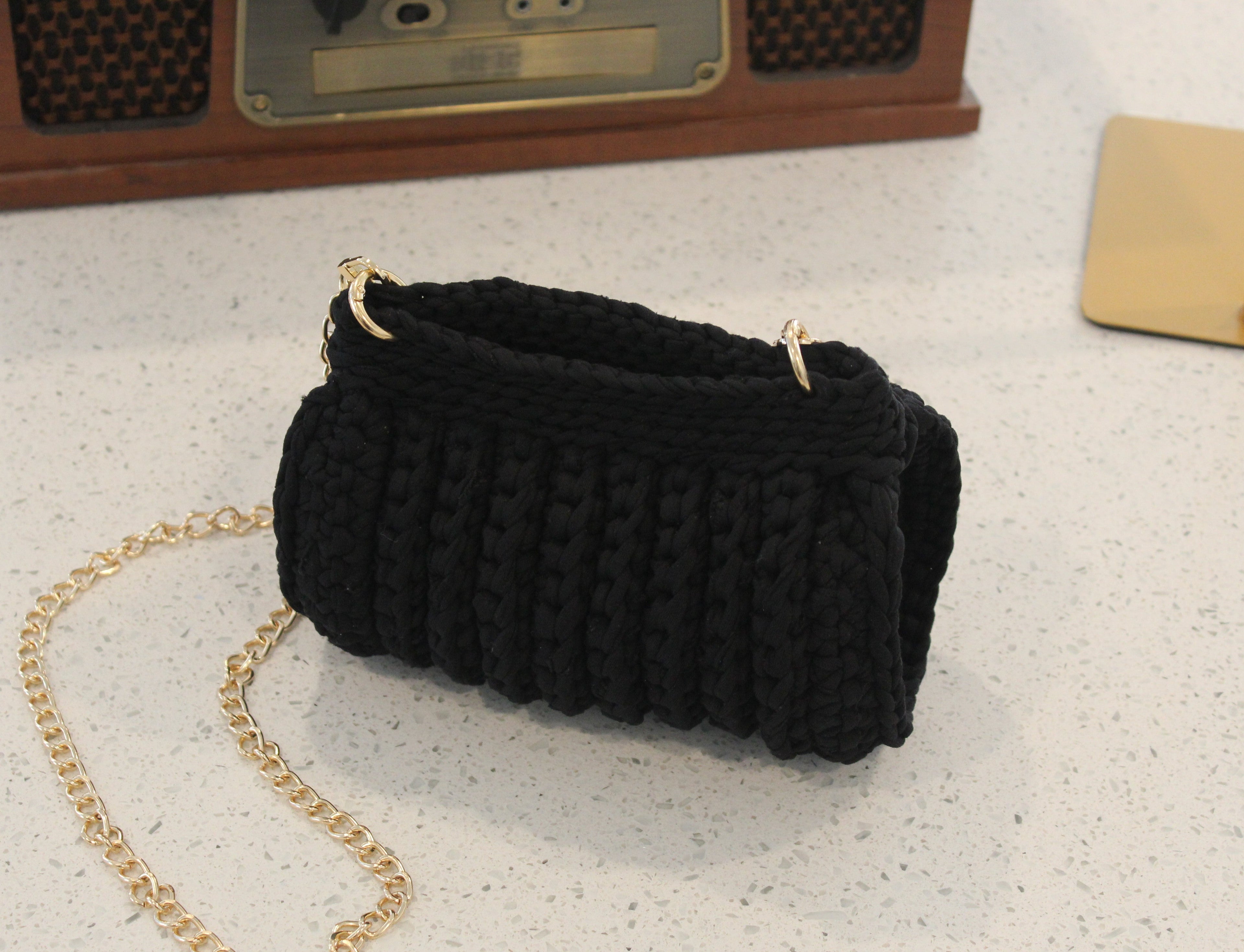 Black hand knitted handbag with a gold chain on a light gray surface.