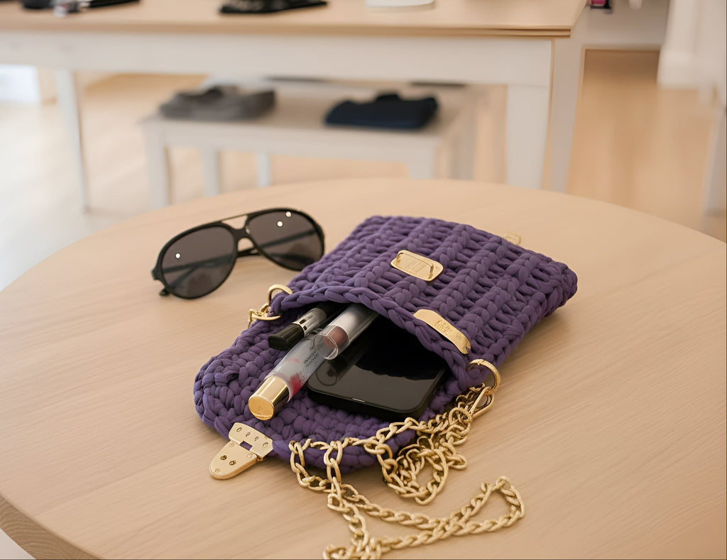 Purple handknitted andbag with gold chain on a wooden table, containing sunglasses and makeup items.